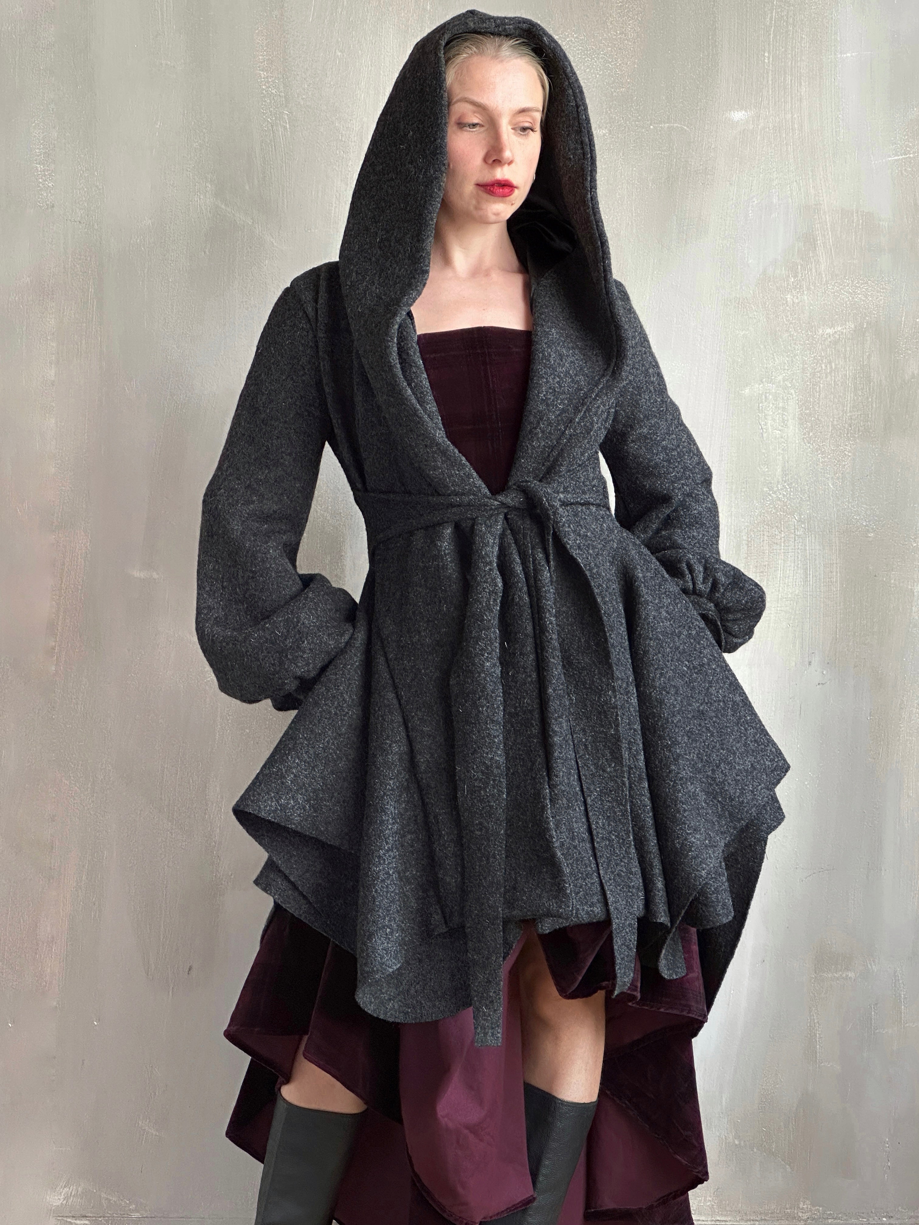 Woman wearing a dark gray hooded coat with a flared silhouette over a burgundy dress against a plain background