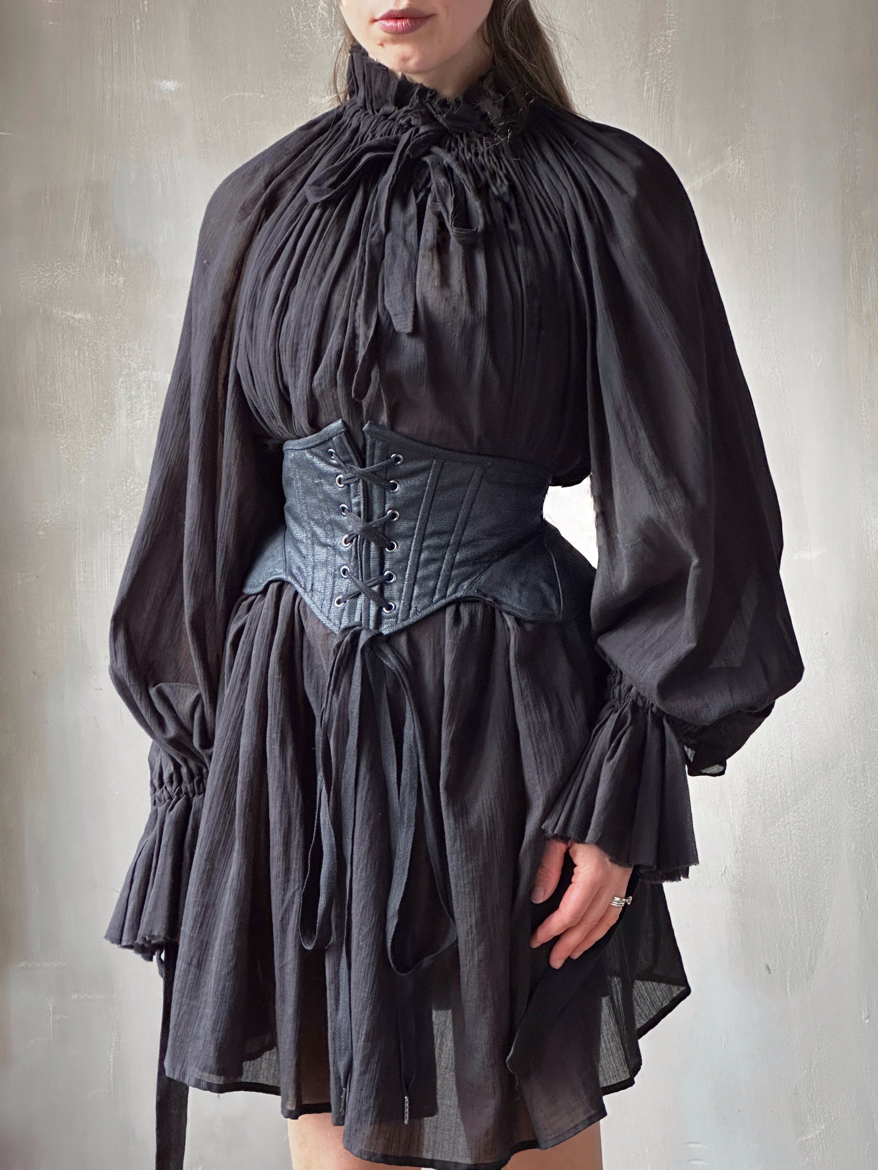 Person wearing a dark elizabethan gothic-style dress with a shiny black corset belt against a plain background